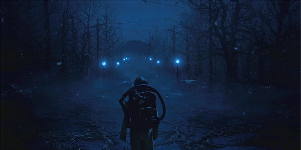Will Byers in the weird blue light of the Upside Down, looking down a desolate street lined with bare trees.