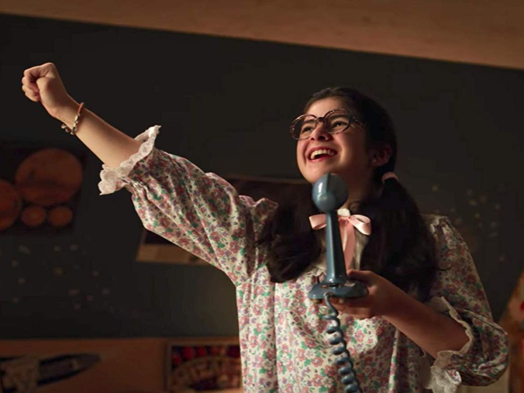 Gabriella Pizzolo as Suzy, smiling as she sings into a ham radio microphone, one arm raised in triumph.