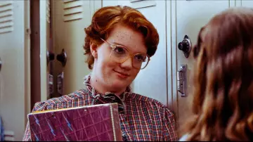 Shannon Purser as Barb Holland, standing by a locker and holding a Trapper Keeper binder.