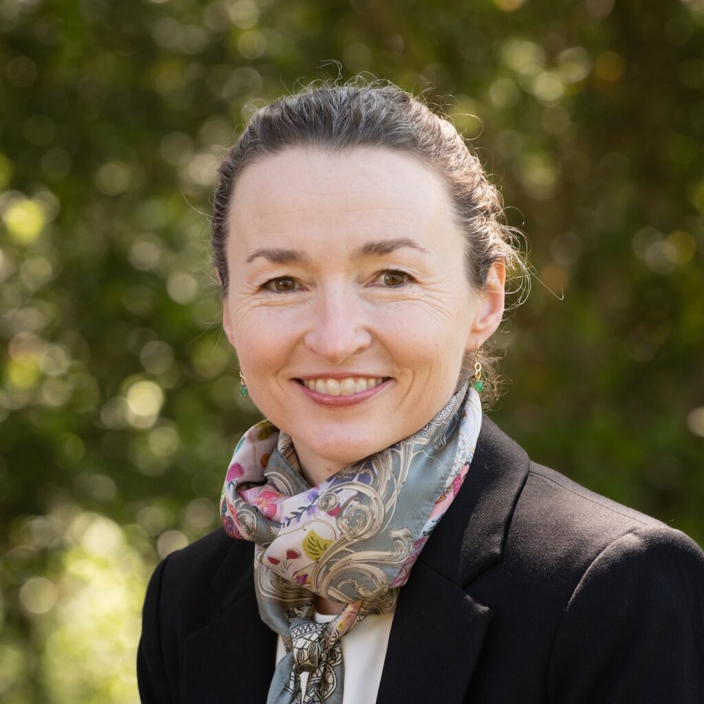 A headshot of Joanna Gardner who is smiling because there are leafy green trees in the background.