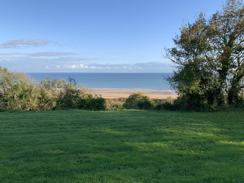 A landscape view of a beach in Normandy with green grass in the foreground, then shrubs and trees, then a stretch of sand, then the blue ocean with clouds off in the distance at the horizon and a big blue sky overhead.